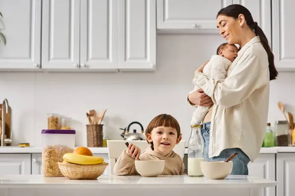 Freudige attraktive Mutter posiert beim Frühstück neben ihrem kleinen Sohn mit Neugeborenem in den Händen — Stockfoto