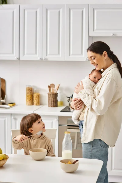 Freudig attraktive Mutter posiert neben ihrem kleinen Sohn mit Neugeborenem in den Händen beim Frühstück — Stockfoto