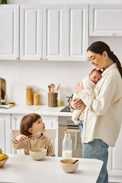 Joyful attractive mother posing next to her toddler son with newborn in hands during breakfast — Stock Photo