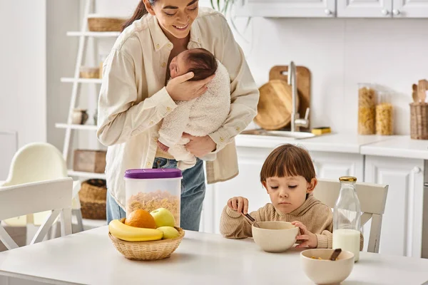 Jolly attractive mother posing next to her toddler son with newborn baby in hands during breakfast — Stock Photo