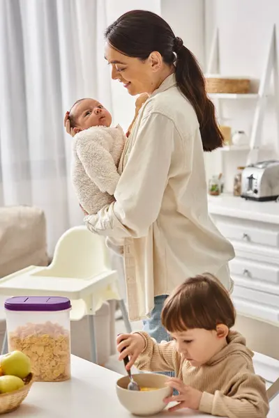 Feliz atraente mãe posando ao lado de seu filho criança com recém-nascido em mãos durante o café da manhã — Fotografia de Stock