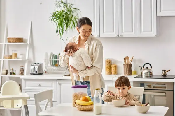 Cheerful attractive mother posing next to her toddler son with newborn in hands during breakfast — Stock Photo