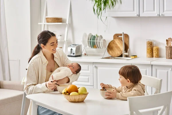 Joyful beautiful mother posing next to her toddler son with newborn in hands during breakfast — Stock Photo