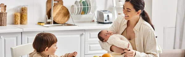 Jolly beautiful mother posing next to her toddler son with newborn in hands during breakfast, banner — Stock Photo