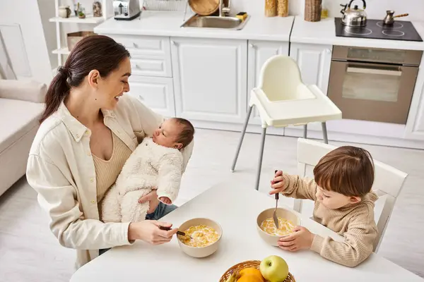 Soins belle mère avoir le petit déjeuner avec son fils tout-petit et tenant son nouveau-né — Photo de stock