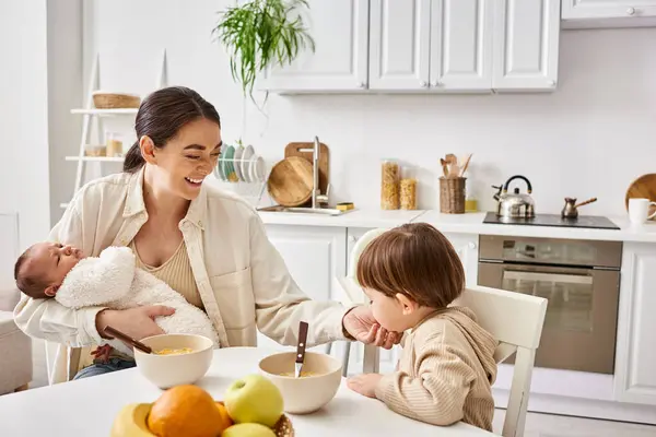 Loving beautiful mother having breakfast with her toddler son and holding her newborn baby — Stock Photo