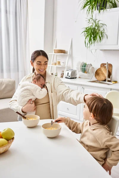 Loving attractive mother having breakfast with her toddler son and holding her newborn baby — Stock Photo