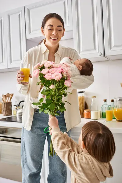 Adorable tout-petit garçon donnant des fleurs à sa belle mère joyeuse avec son frère nouveau-né dans les mains — Photo de stock