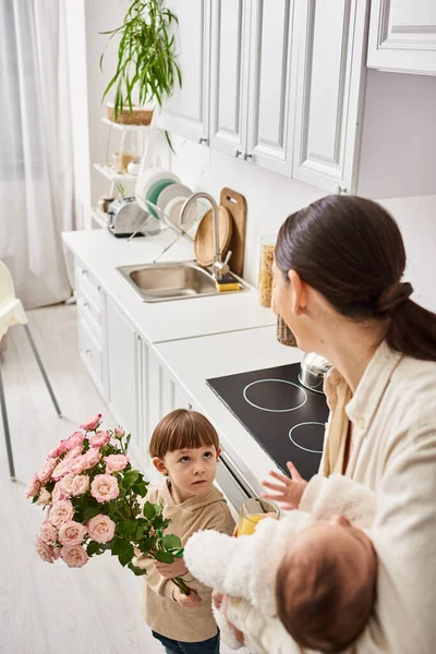 Adorable tout-petit garçon donnant des fleurs à sa belle mère joyeuse avec son frère nouveau-né dans les mains — Photo de stock