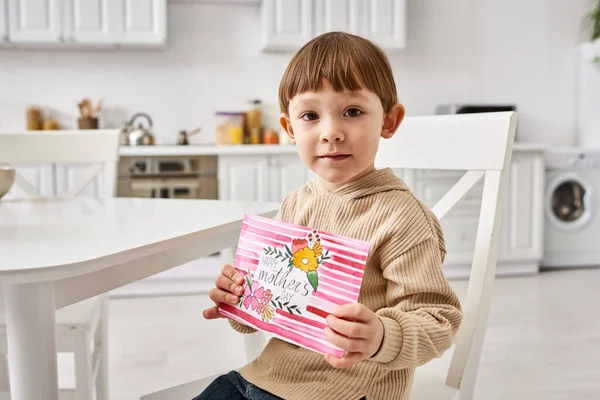 Adorable little boy in comfortable attire sitting and holding greeting card on Mothers day — Stock Photo