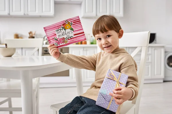 Adorable lindo niño en acogedora ropa de casa posando con presente y tarjeta de felicitación en el día de las madres - foto de stock