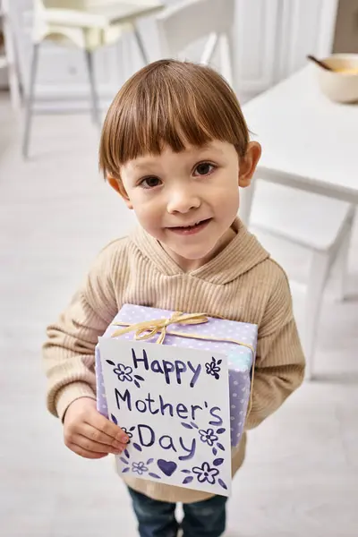 Adorable lindo niño en ropa de casa casual posando con presente y tarjeta de felicitación en el día de las madres - foto de stock