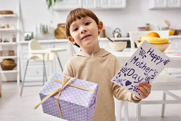 Joyous cute little boy in casual attire posing with present and flowers in his hands, Mothers day — Stock Photo