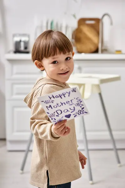 Adorable niño alegre posando con tarjeta de felicitación para el día de las madres y sonriendo a la cámara - foto de stock