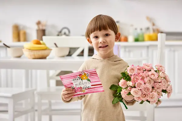 Alegre lindo niño pequeño en traje casual posando con flores ramo y tarjeta de felicitación, día de las madres - foto de stock