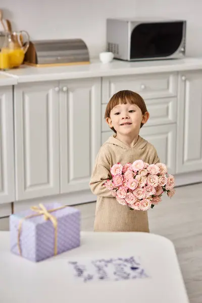 Menino bonito criança em homewear segurando buquê de flores perto da mesa com presente e cartão postal nele — Fotografia de Stock