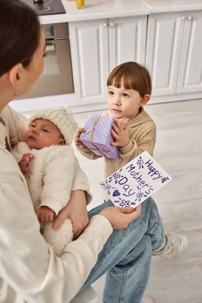 Adorable boy giving gift and postcard to his mom while she holding his newborn brother, Mothers day — Stock Photo