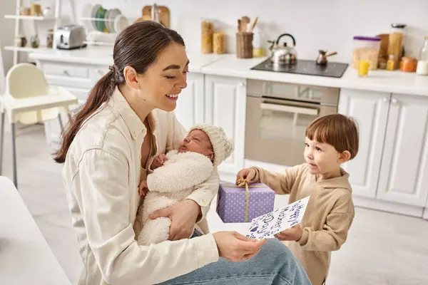 Toddler boy giving gift and postcard to his mom while she holding his newborn brother, Mothers day — Stock Photo