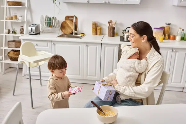 Cute boy giving present and postcard to his mom while she holding his newborn brother, Mothers day — Stock Photo
