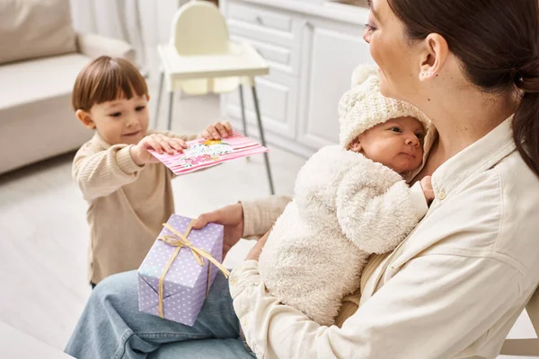 Adorable boy giving present and postcard to mom while she holding his newborn brother, Mothers day — Stock Photo