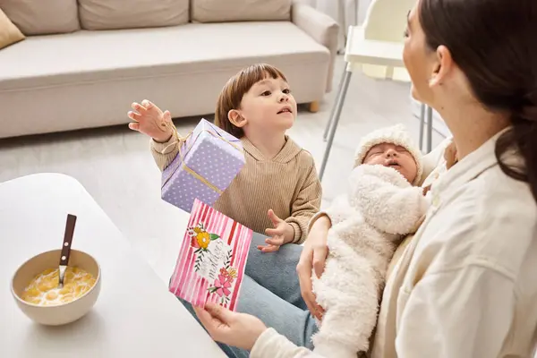 Toddler boy giving present and postcard to mom while she holding his newborn brother, Mothers day — Stock Photo