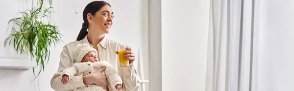 Alegre madre amorosa sosteniendo a su bebé recién nacido y sosteniendo jugo de naranja, crianza moderna, pancarta - foto de stock