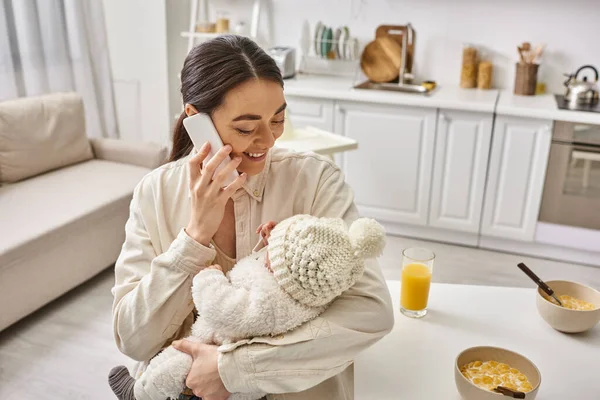 Attractive mother in homewear holding warmly her baby boy and talking by phone, modern parenting — Stock Photo