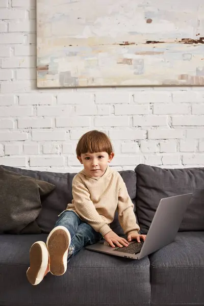 Adorable mignon tout-petit garçon en vêtements confortables assis sur le canapé avec ordinateur portable et regardant la caméra — Photo de stock