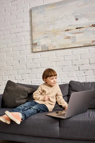 Concentrated adorable cute toddler boy in cozy homewear sitting on sofa and looking at laptop — Stock Photo