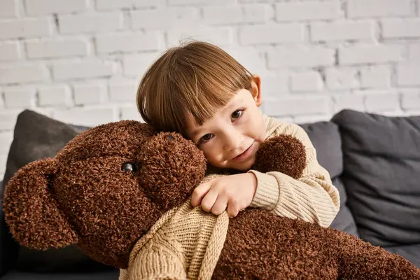 Jolly little boy in homewear hugging his teddy bear while sitting on couch and looking at camera — Stock Photo