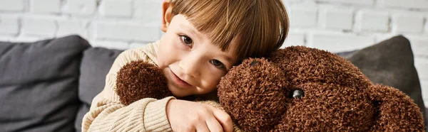 Happy little boy hugging his teddy bear while sitting on couch and looking at camera, banner — Stock Photo