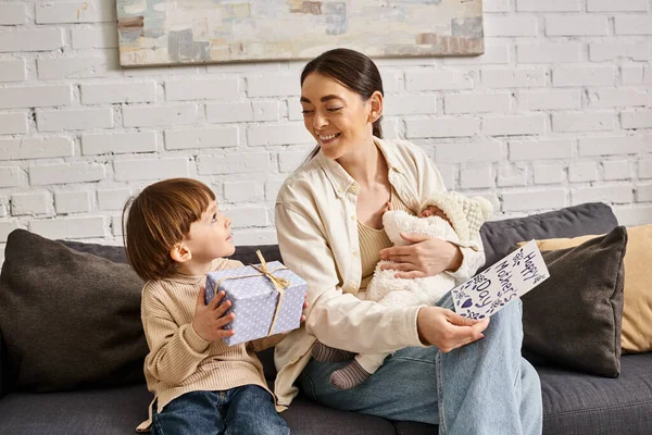 Atractiva mujer sentada en el sofá con sus hijos con presente y tarjeta de felicitación en el día de la madre - foto de stock