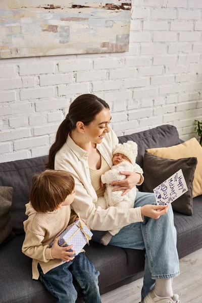 Gioiosa bella donna seduta sul divano con i suoi figli con regalo e biglietto di auguri il giorno della mamma — Foto stock