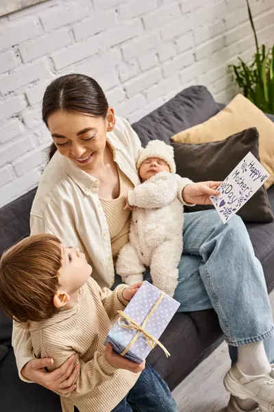 Atractiva mujer sentada en el sofá con sus hijos con presente y tarjeta de felicitación en el día de la madre - foto de stock