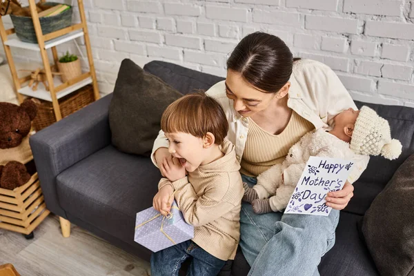 Cheerful woman sitting on sofa with her sons with present and greeting card on Mothers day — Stock Photo
