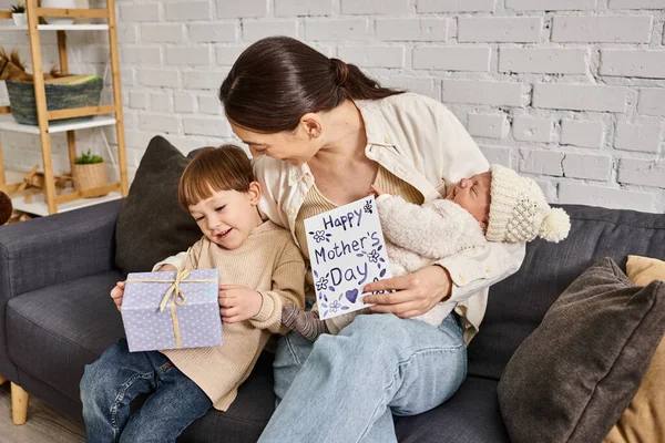 Belle femme assise sur le canapé avec ses fils avec cadeau et carte de vœux sur la fête des mères — Photo de stock