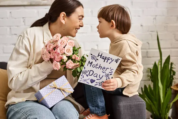 Гарна весела мати позує на дивані зі своїм маленьким сином з квітковим букетом і подарунком, День матері — стокове фото