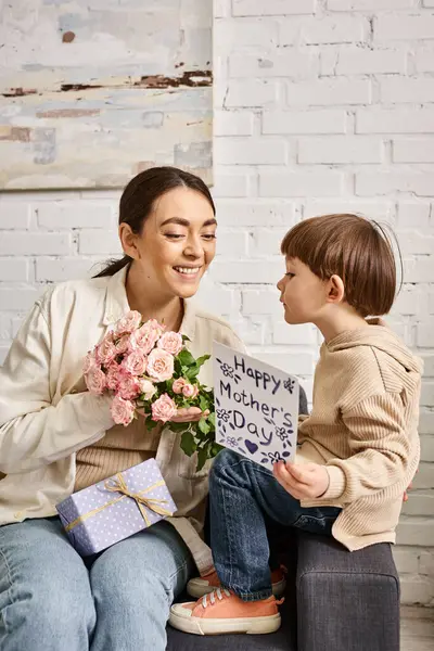 Amorevole madre allegra in posa sul divano con il suo bambino figlio con mazzo di fiori e presente, Festa della mamma — Foto stock