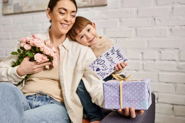 Досить щаслива мати позує на дивані зі своїм маленьким сином з квітковим букетом і подарунком, День матері — стокове фото