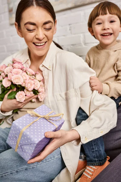 Любляча щаслива мати позує на дивані зі своїм маленьким сином з квітковим букетом і подарунком, День матері — стокове фото