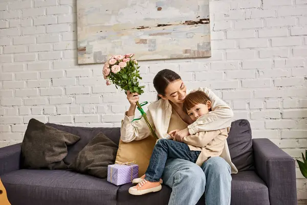 Mère heureuse attentionnée posant sur le canapé avec son fils tout-petit avec bouquet de fleurs et présent, Fête des mères — Photo de stock