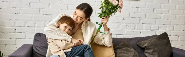 Belle mère posant sur canapé avec son mignon fils tout-petit avec bouquet de fleurs, fête des mères, bannière — Photo de stock