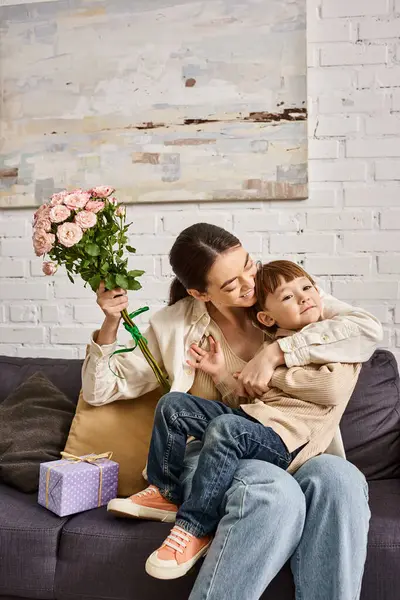 Bella madre allegra posa sul divano con il suo bambino figlio con mazzo di fiori e presente, Festa della mamma — Foto stock