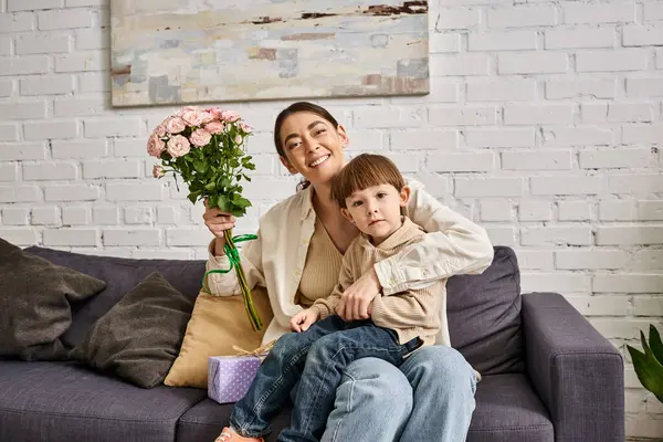 Aimante mère joyeuse posant sur le canapé avec son fils tout-petit avec bouquet de fleurs et présent, Fête des mères — Photo de stock