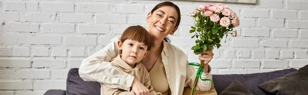 Belle mère posant sur le canapé avec son mignon fils tout-petit avec bouquet de fleurs, fête des mères, bannière — Photo de stock