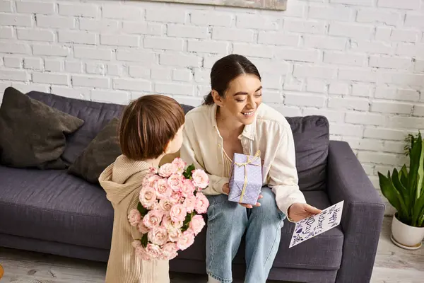 Cute toddler boy giving his jolly mom present and postcard with flower bouquet on Mothers day — Stock Photo