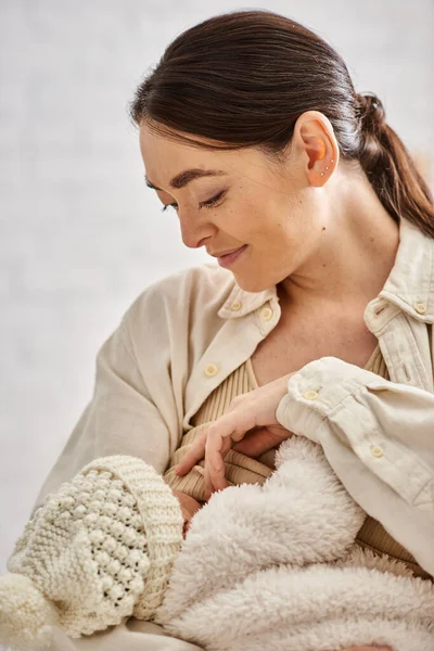 Ansprechende fürsorgliche, fröhliche Mutter in kuscheliger Hauskleidung, die ihr neugeborenes Kind stillt, moderne Elternschaft — Stockfoto