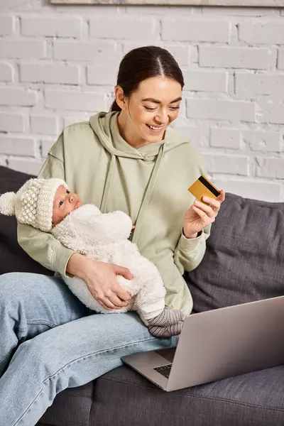 Atractiva madre alegre en ropa de casa sosteniendo a su bebé en la fuente de la computadora portátil con tarjeta de crédito en la mano - foto de stock