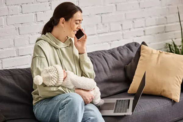Mujer atractiva alegre en traje acogedor mirando el ordenador portátil y sosteniendo a su bebé mientras habla por teléfono - foto de stock