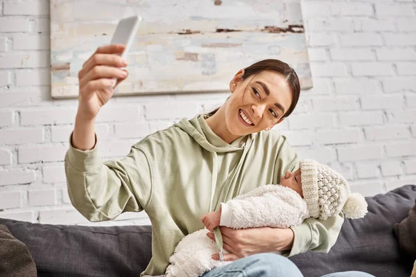 Fröhliche schöne Mutter in lässiger Kleidung macht Selfie, während sie ihren neugeborenen Jungen hält — Stockfoto